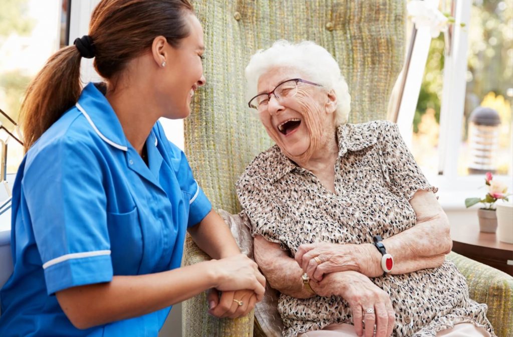 A caregiver kneeling beside a sitting older adult in memory care laughing and smiling together.