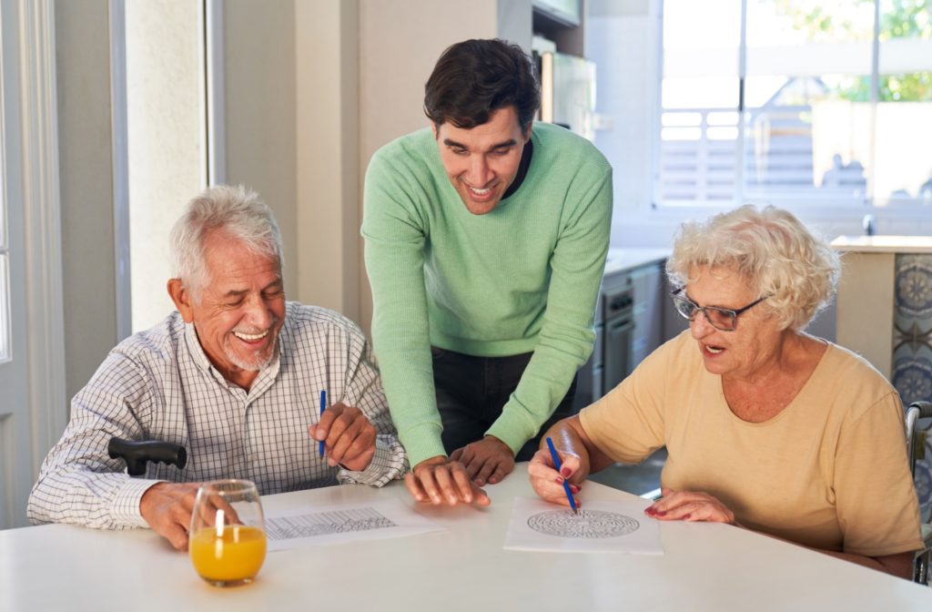 A caregiver in a green shirt helps two seniors with dementia complete puzzles to aid their memory