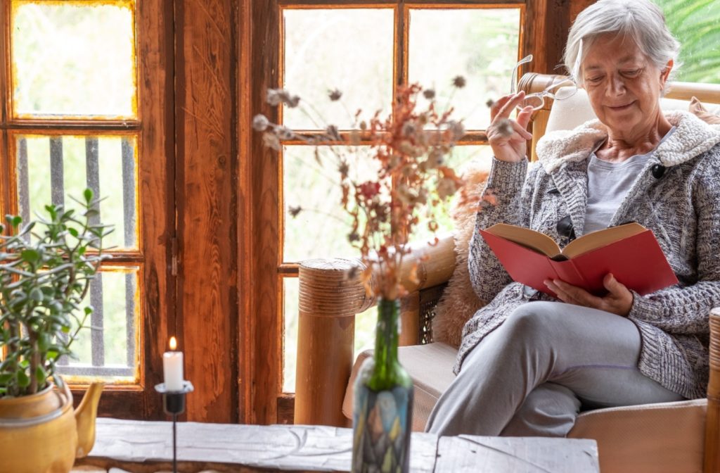 An older woman relaxing indoors, reading a book near a cozy window with natural light and a candle nearby.