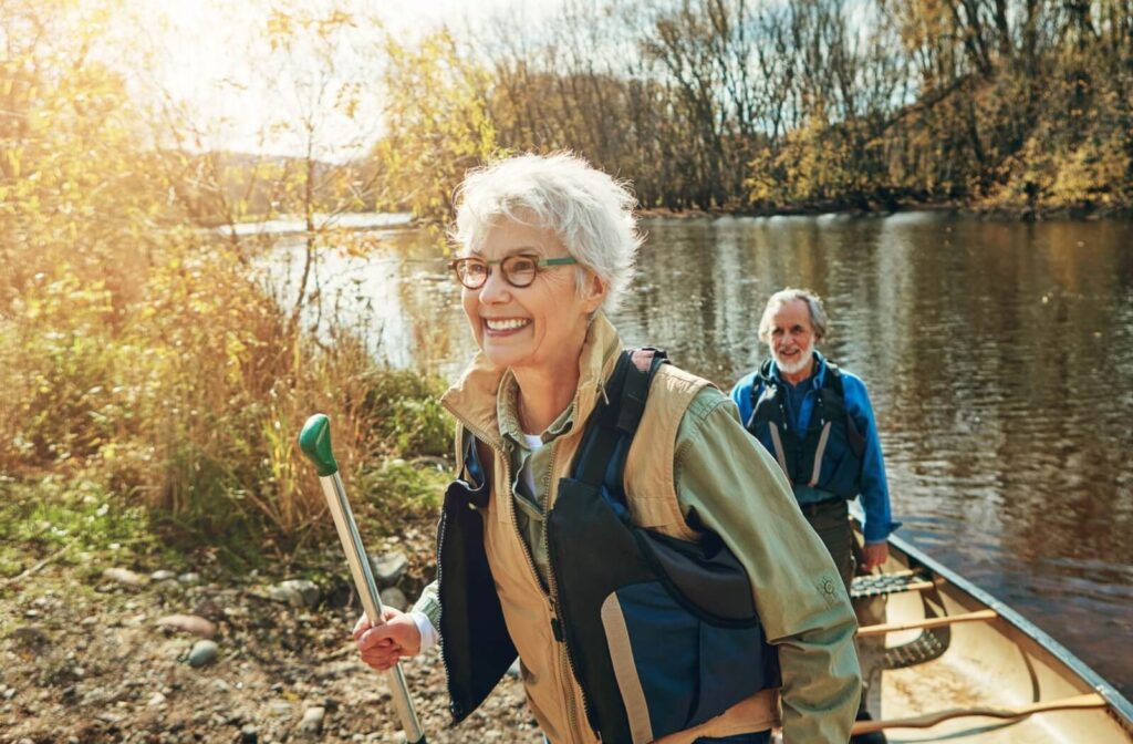 Smiling older adult in life vest carrying paddle with companion exiting canoe near riverbank under autumn trees