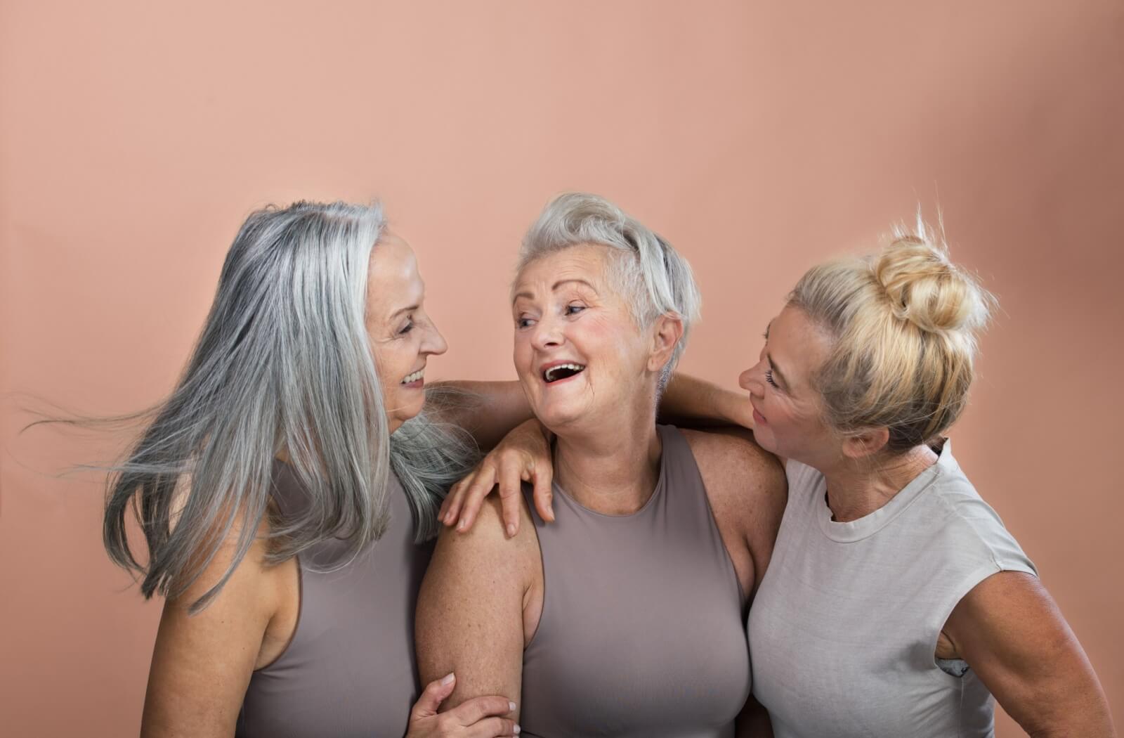 Against a pale pink background, three seniors in neutral colours smile and embrace each other and the beauty of their age