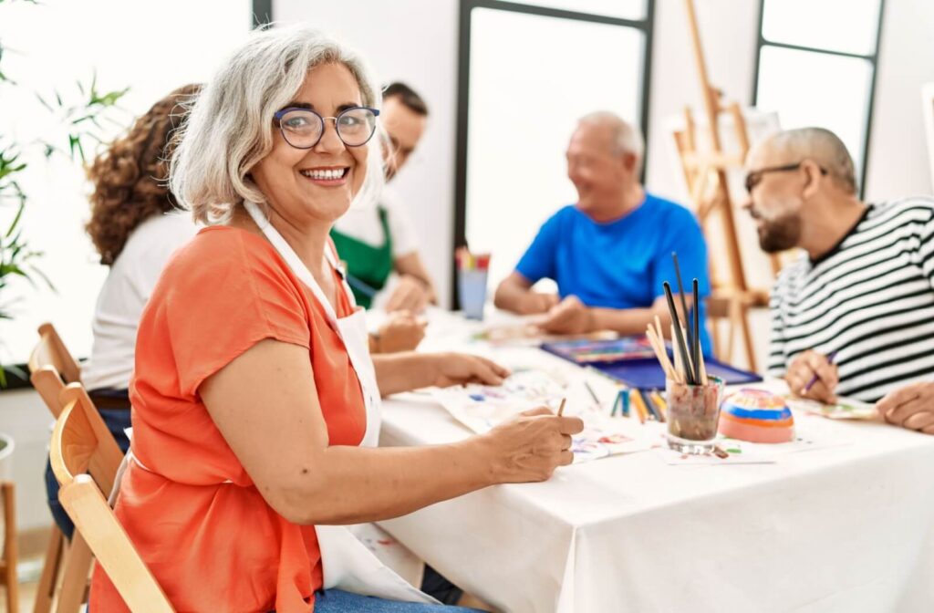 A smiling senior poses with a paintbrush in hand, during an art class offered by their senior living community