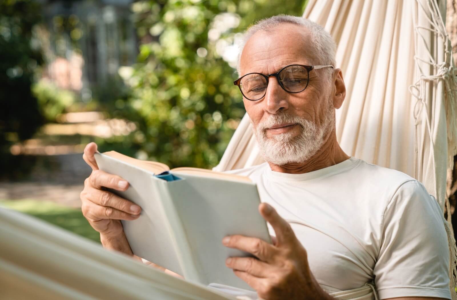 A senior reads a hardcover book while relaxing in a hammock as a way to improve their memory
