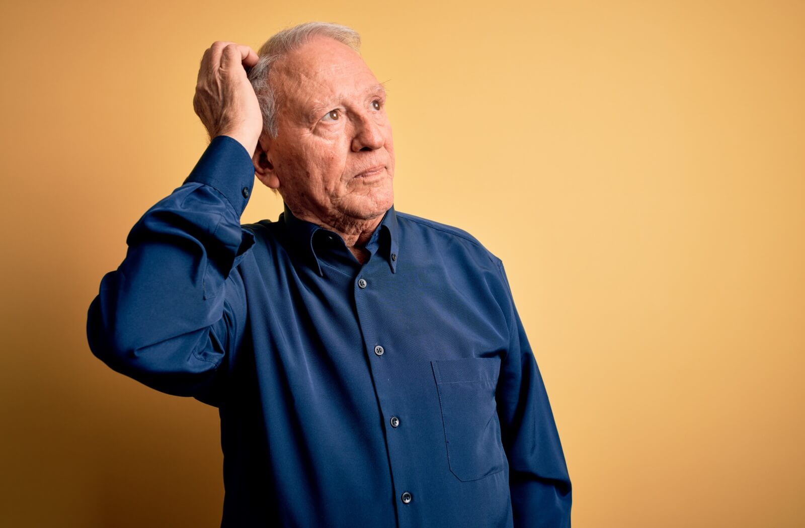 Against a yellow backdrop, a senior in a blue button-down shirt scratches their head in confusion while looking to the side